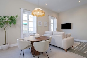 Een moderne woonkamer met een ronde eettafel, witte stoelen, een witte bank, een potplant en een tv aan de muur.