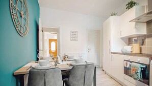 Une cuisine moderne avec un coin repas, des meubles blancs, une horloge et un panneau décoratif au mur.