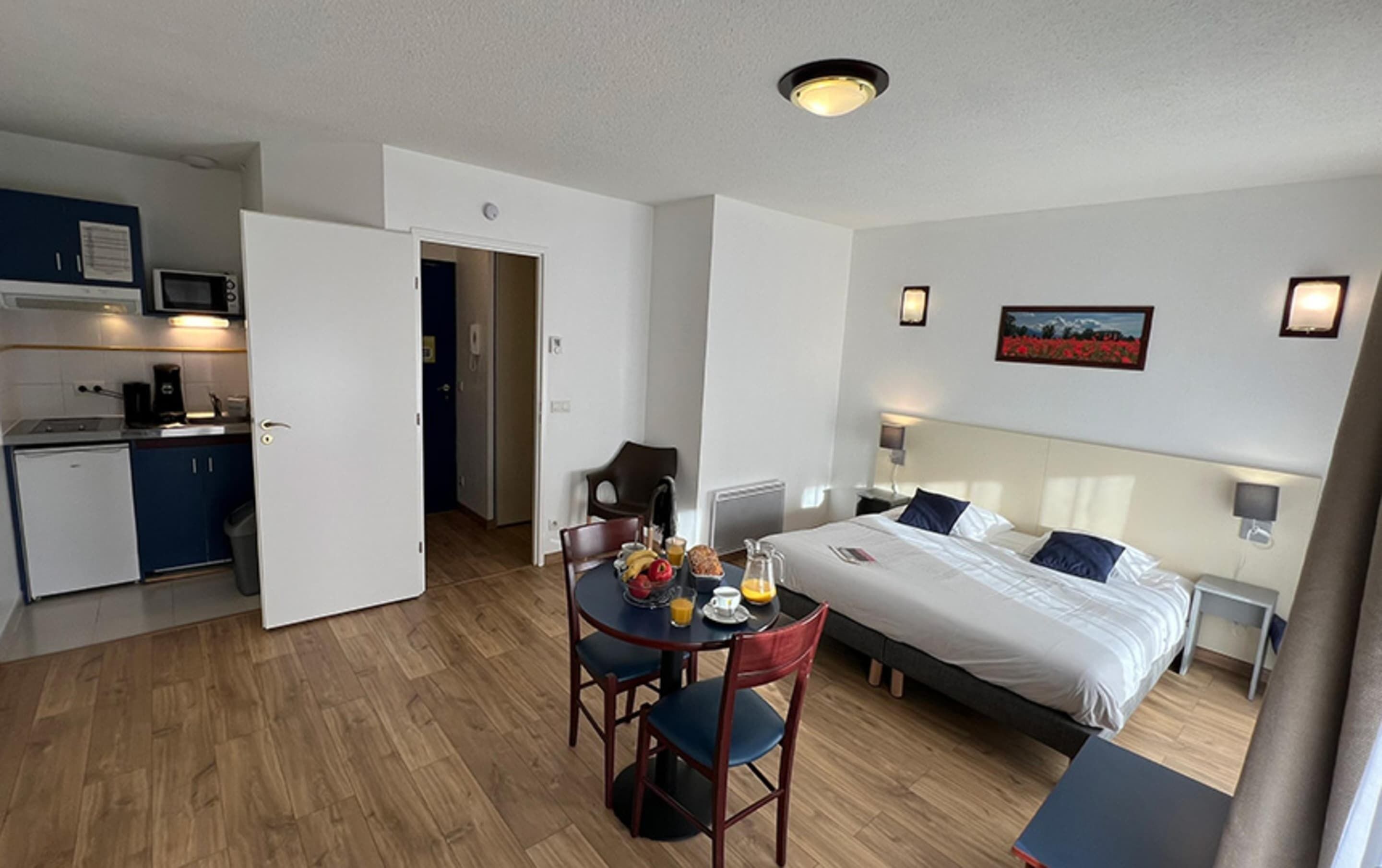 A hotel room with a bed, bedside tables, a desk with a chair, a dining table with chairs, a kitchen area with a microwave, and a door leading to another room.
