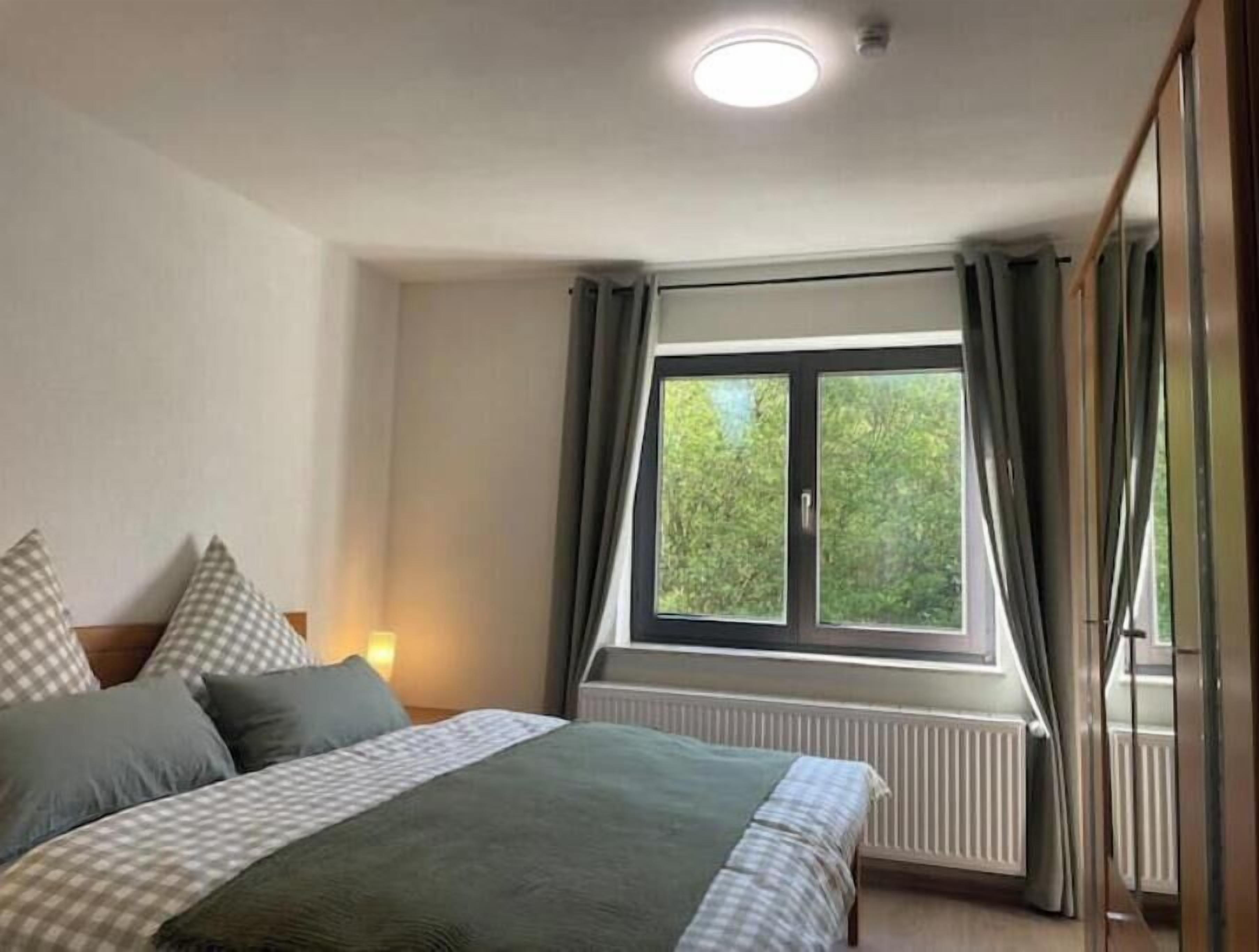 Un dormitorio con cama, ventana con cortinas, radiador y lámpara en el techo.