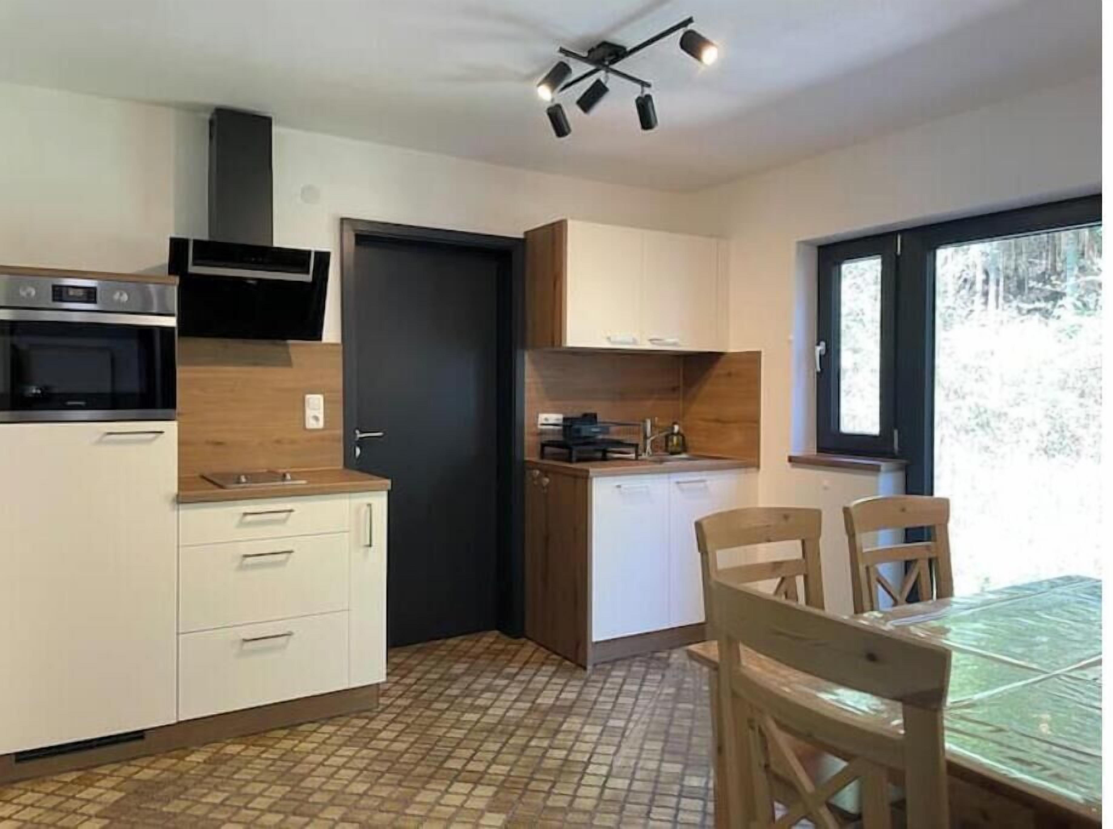 Een kleine keuken met een kookplaat, oven en magnetron, een eettafel met stoelen, en een raam met uitzicht op bomen.