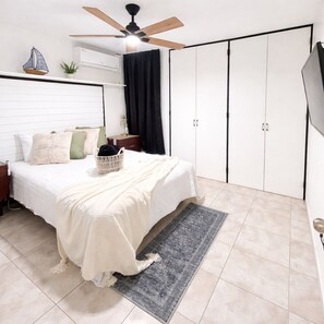 Ein Schlafzimmer mit einem Bett, einem Deckenventilator, einem Fernseher, einem Kleiderschrank und einer kleinen Pflanze.