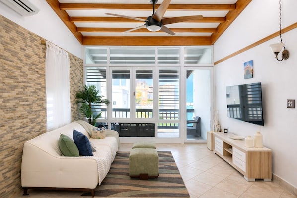 Ein Wohnzimmer mit einem weißen Sofa, einem grünen Hocker, einem Deckenventilator, einem Fernseher und einem großen Fenster mit Blick ins Freie.