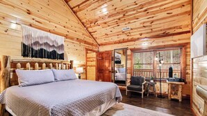 Une chambre dans un chalet en rondins, avec un grand lit, un plafond en bois, une fenêtre avec des stores et une petite table accompagnée d’une chaise.