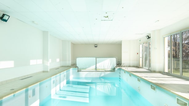 En stor indendørs swimmingpool med krystalklart vand, flisegulv og udsigt til det fri gennem store vinduer.