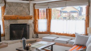 A cozy living room with a stone fireplace, wooden beams, a seating area with cushions, and a view through large windows. - NOTRO 2 Tero (San Carlos de Bariloche)