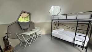 Camera con letto a castello, un piccolo tavolo e sedie, una finestra vista su vegetazione e un'originale lampada a forma esagonale.