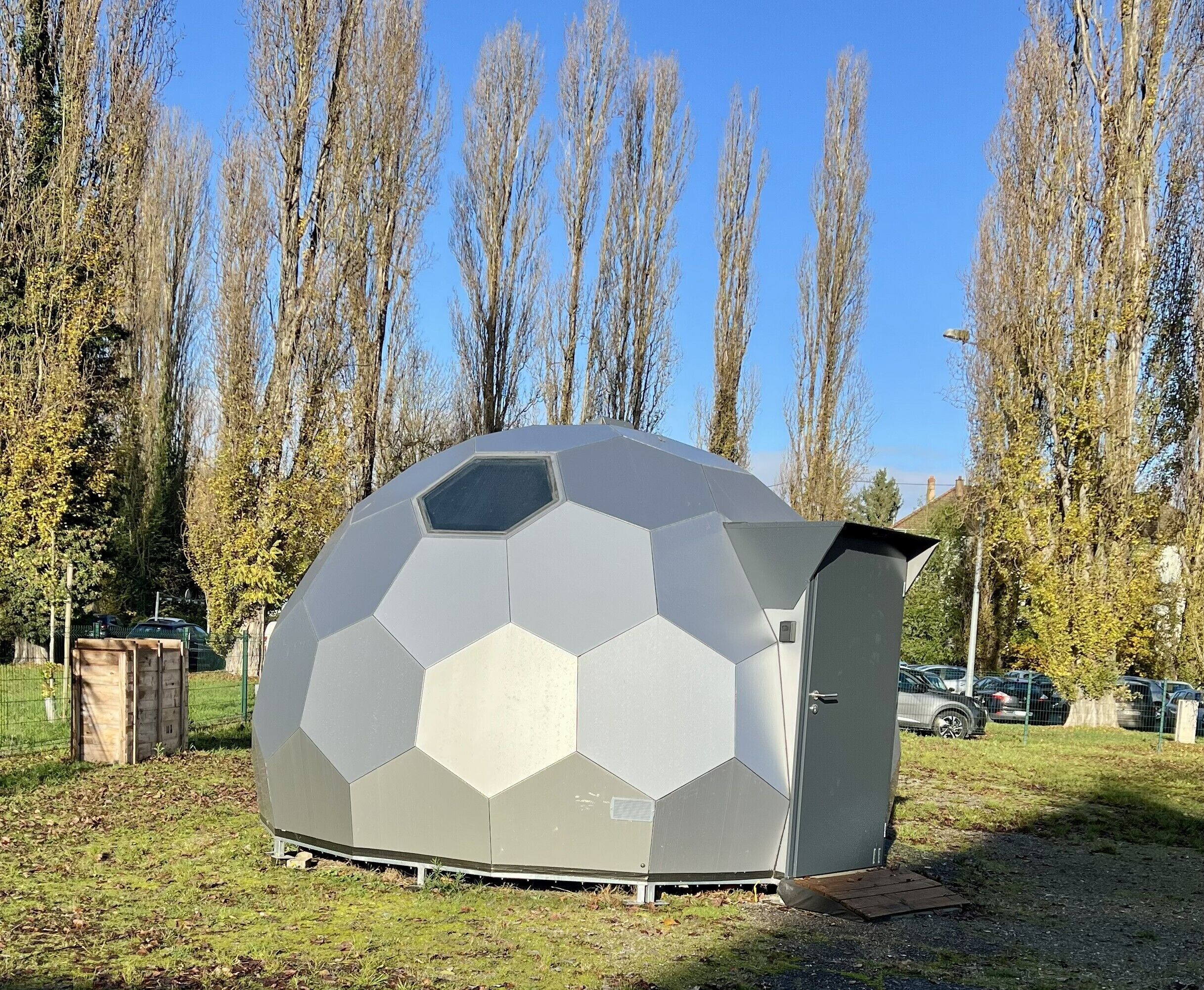 Una estructura hexagonal que recuerda a un balón de fútbol, con una puerta y una pequeña ventana, ubicada en un área cubierta de césped y con árboles al fondo.