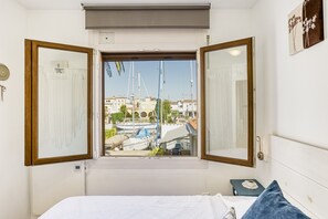 Ein Schlafzimmer mit großem Fenster, einem Bett, einem Nachttisch und Blick auf Boote und Gebäude.