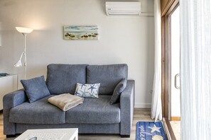 Ein graues Sofa mit Decke und Kissen mit Sternenmuster, eine weiße Stehlampe, ein Kunstwerk zum Thema Strand und eine blaue Matte mit Meeresschnecken und dem Wort 'welcome' auf Holzdielen.