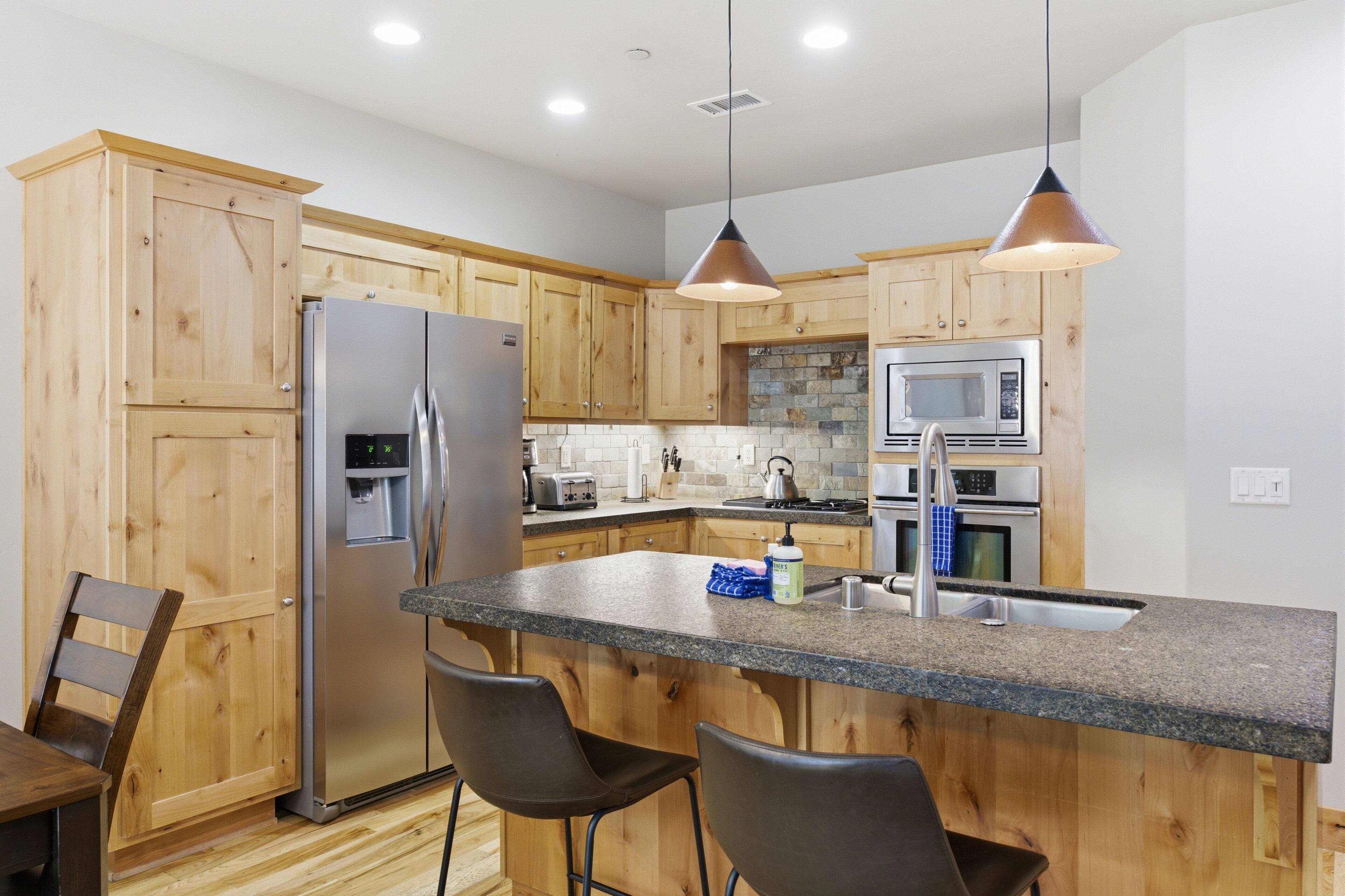 Una cocina moderna con gabinetes de madera, electrodomésticos de acero inoxidable y encimeras de granito, además de taburetes de bar.
