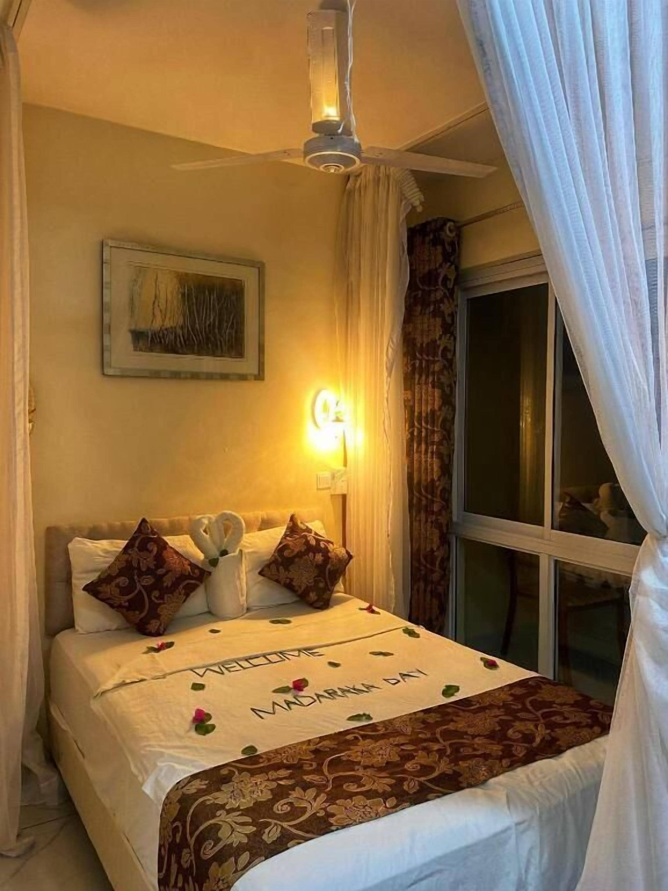 Habitación de hotel con cama, ventilador de techo, luz nocturna, ventana con cortinas e imagen enmarcada en la pared.