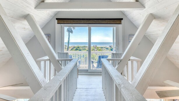 Vista da un balcone con ringhiera in legno, che conduce a una grande porta in vetro e offre una visuale chiara sull'oceano e sulla spiaggia.