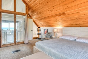 Quarto com teto inclinado, cama, cadeira, luminária e vista para o oceano.