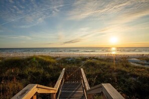 Ein Holzsteg führt zu einem Strand, im Hintergrund ein Sonnenuntergang.