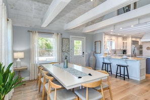 A spacious dining area with a long table, several chairs, a vase with flowers, and a view of the kitchen. - Seaside Siesta - Ocean Front, Private Beach Walkover, Charming (St. Augustine)