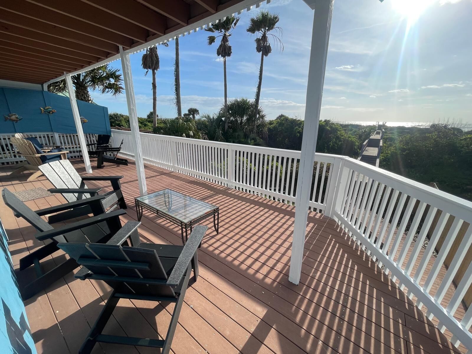 A covered deck with outdoor seating, a glass table, and a view of palm trees and the ocean.
