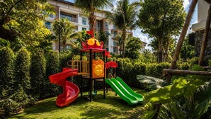 Un’area giochi con scivoli e strutture di arrampicata, circondata da vegetazione lussureggiante e un edificio sullo sfondo.