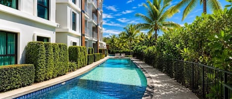 Una piscina junto a un edificio de varios pisos con balcones, rodeada de setos cuidados y palmeras.