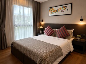 A hotel room with a large bed, a nightstand, a wall-mounted lamp, and a window with curtains. - Modern 1br Apartment in Rawai | 2min to beach (Rawai)