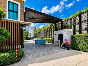 A modern building entrance with a sign, a paved pathway, and a motorbike with a delivery box. - Cozy 1br in Rawai Quiet Area balcony&fast wi-fi (Rawai)