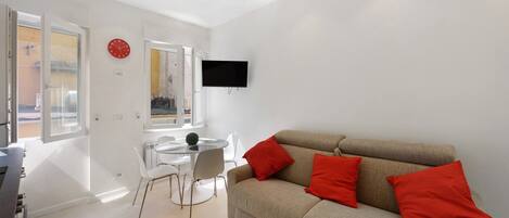 A modern living room with a sofa, red cushions, a flat-screen TV, a dining table with chairs, and a window.