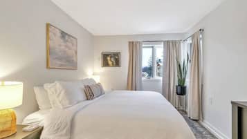 A bedroom with a bed, a nightstand, a lamp, a window with curtains, and a plant.