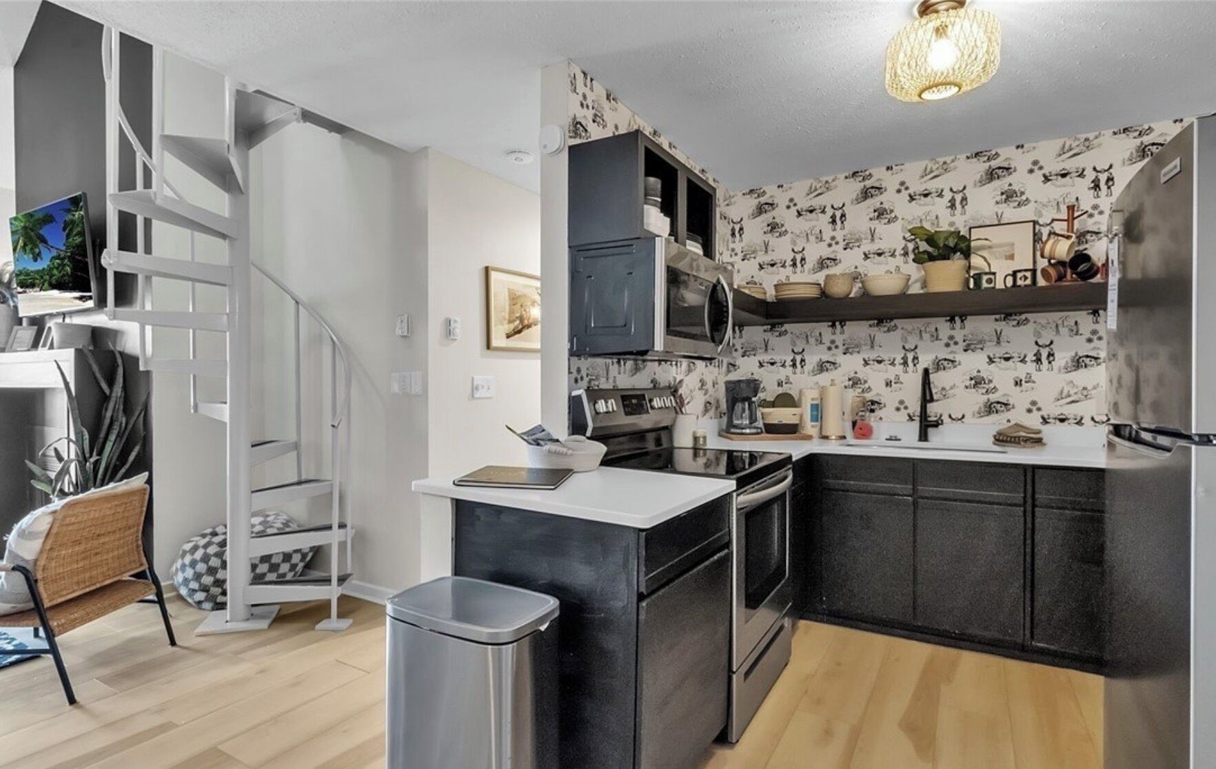 A modern kitchen with a built-in stove, microwave, and black cabinets. There is a small island with a sink and a trash can. The kitchen has a patterned backsplash and a light fixture above the island.