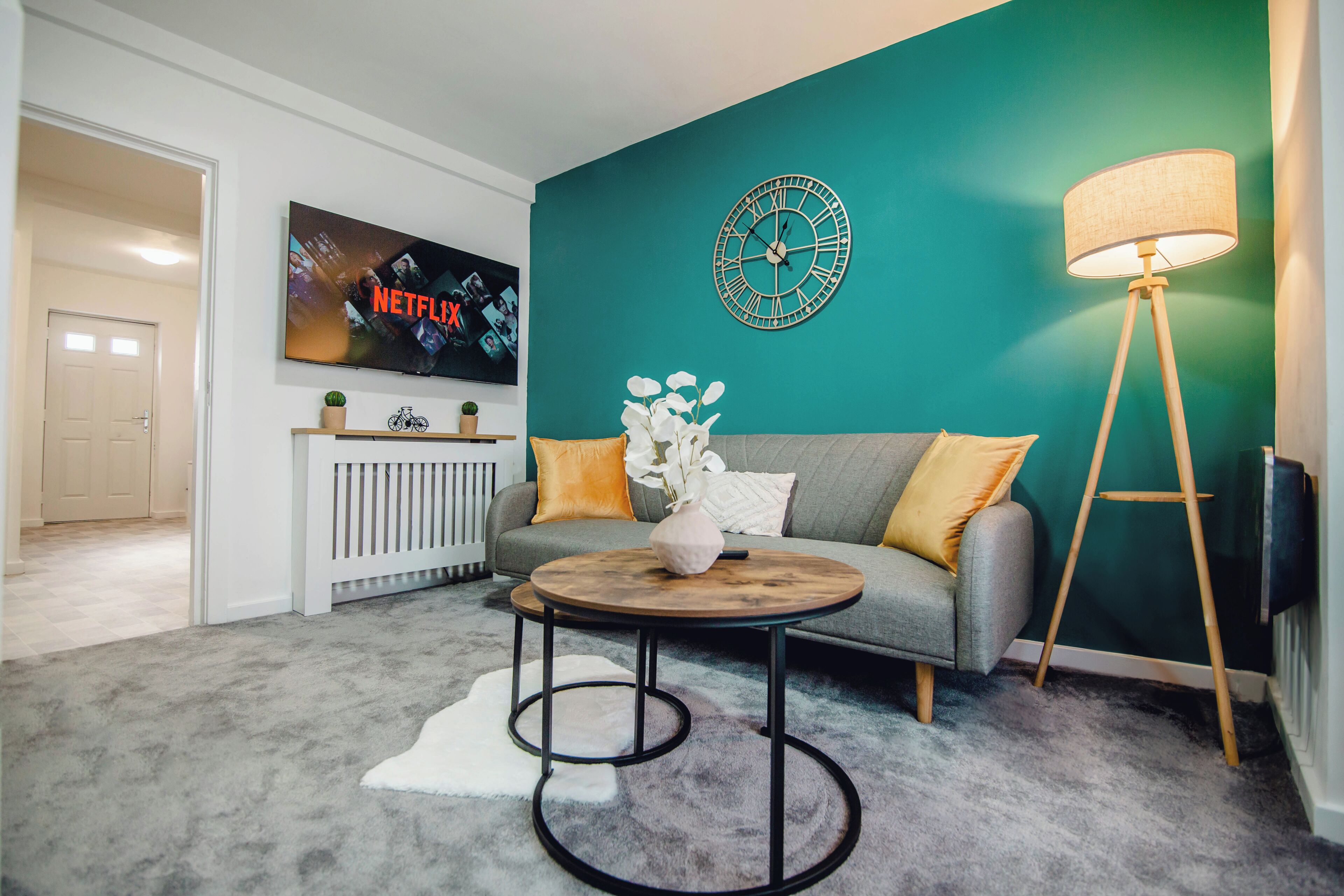 A living room with teal walls, a grey sofa, a round coffee table, a floor lamp, and a TV displaying a Netflix advertisement.