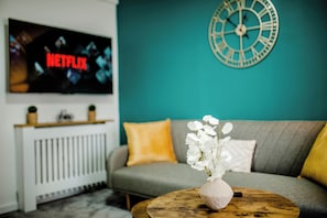 Ein Wohnzimmer mit einer Couch, einem Couchtisch mit einer Vase, einer Wanduhr und einem Fernseher, der 'NETFLIX' anzeigt.