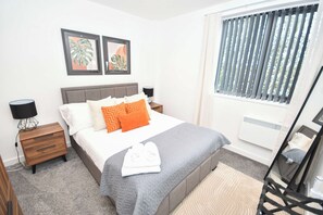 Ein Schlafzimmer mit einem Bett, Nachttischen, einer Lampe, einem Fenster mit Jalousien und einem Bild an der Wand.