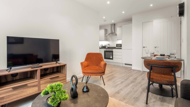 A modern living room with a TV, wooden entertainment unit, orange armchair, dining table with chairs, and a potted plant. - Bright & Spacious 1 Bedroom Apartment in Leeds (Leeds)