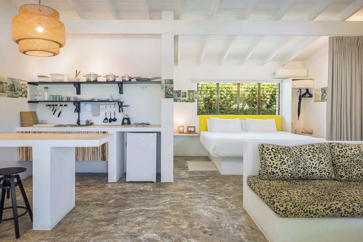 A compact hotel room with a kitchenette, a bed with a yellow headboard, a leopard print sofa, and a large window with a view of greenery.