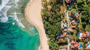 Vista aérea de una zona costera con una playa, un conjunto de edificios con techo rojo y una franja de árboles densa.