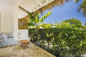 Un área de estar moderna al aire libre con un sofá y una mesa de madera, rodeada de abundante vegetación y un cielo azul despejado.