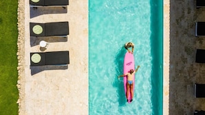 Una piscina con agua turquesa cristalina, una persona sobre un flotador rosa y sillas de descanso negras con cojines verdes.