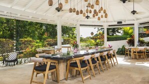 Un espacio comedor al aire libre cubierto, con sillas y mesas de madera, lámparas colgantes y abundante vegetación visible a través del techo transparente.