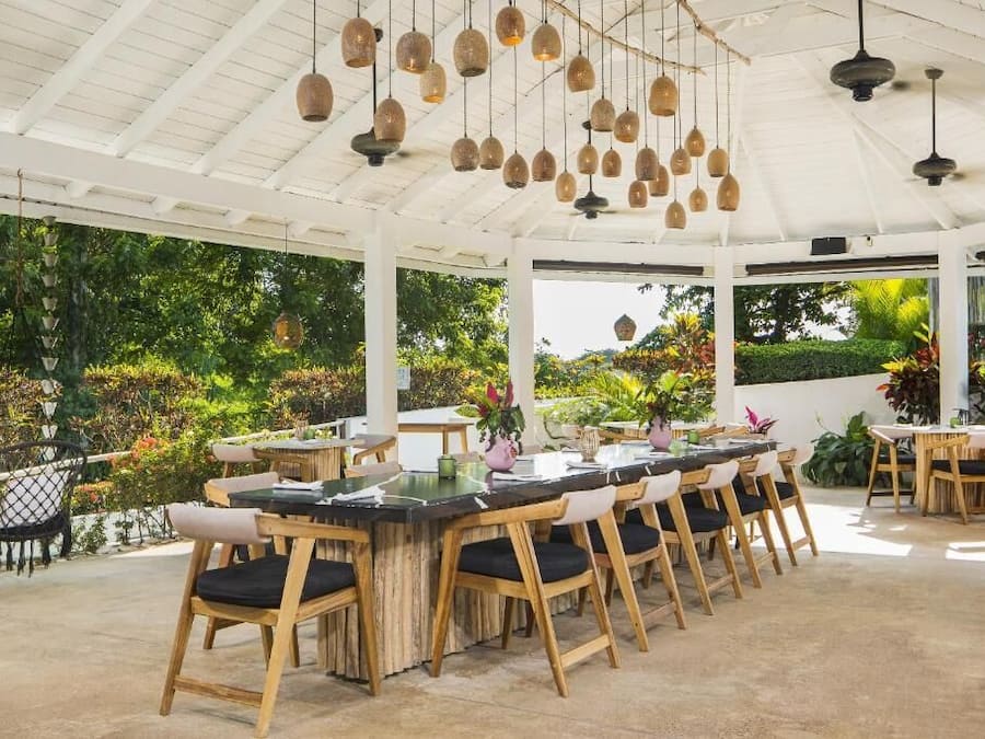 A covered outdoor dining area with wooden chairs and tables, hanging pendant lights, and lush greenery visible through the transparent roof.