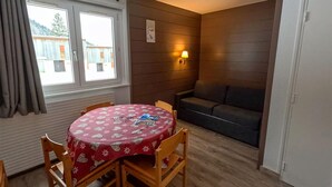 A cozy room with a round table, wooden chairs, a sofa, a window with curtains, and a wall-mounted lamp. - Mountain Flat With Pool in Lelex (Lelex)
