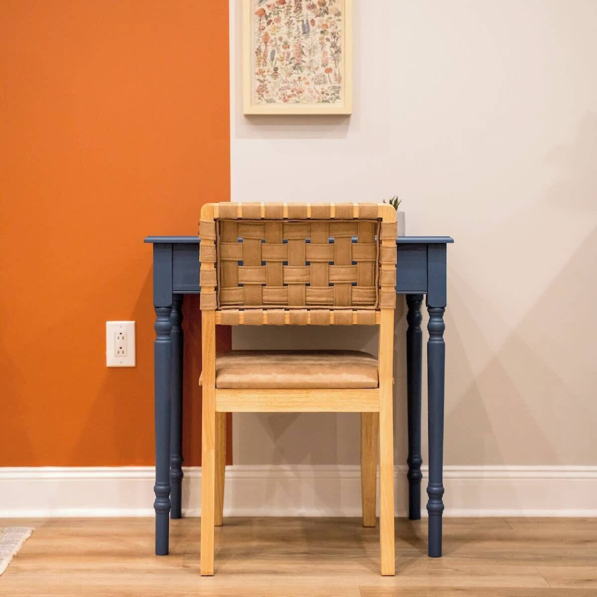 A chair with a woven backrest and a wooden seat, placed between two dark blue tables with turned legs, against an orange wall with a white baseboard.