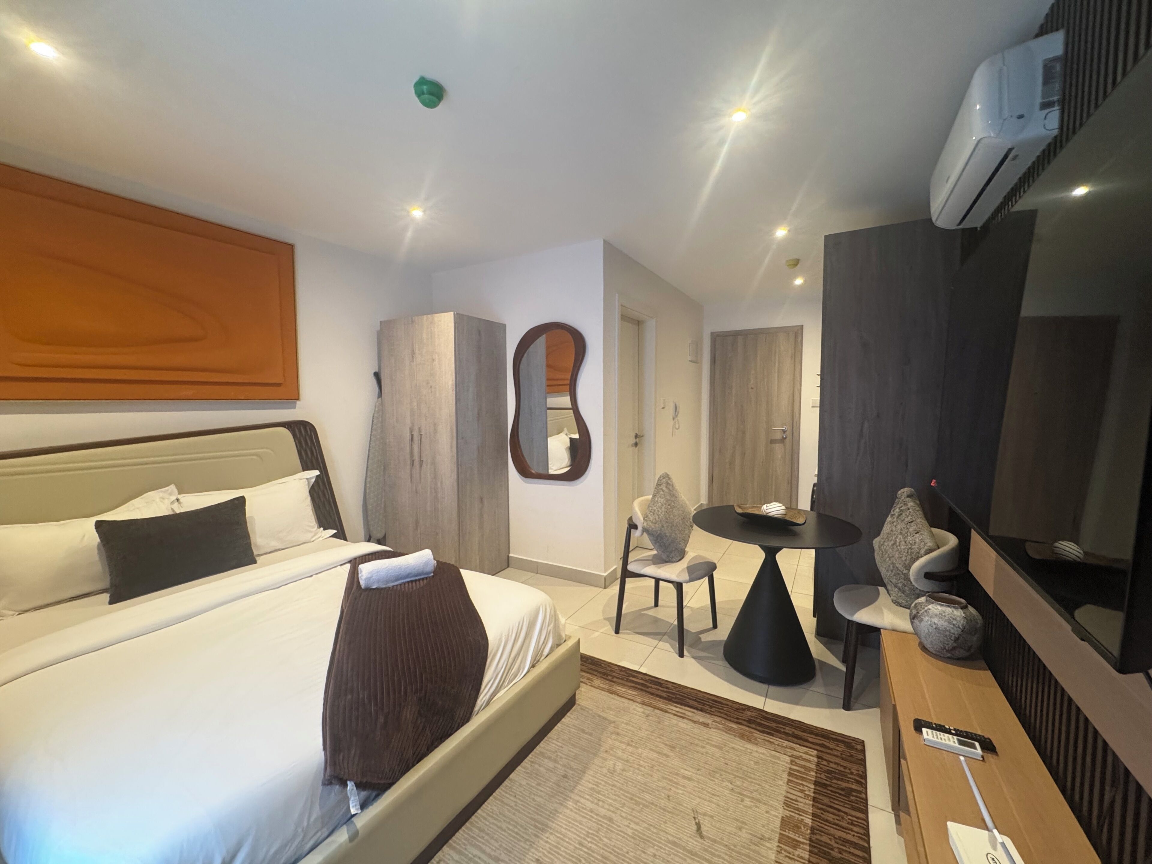 A hotel room with a bed, a desk, a chair, a round table, a mirror, and an air conditioning unit.