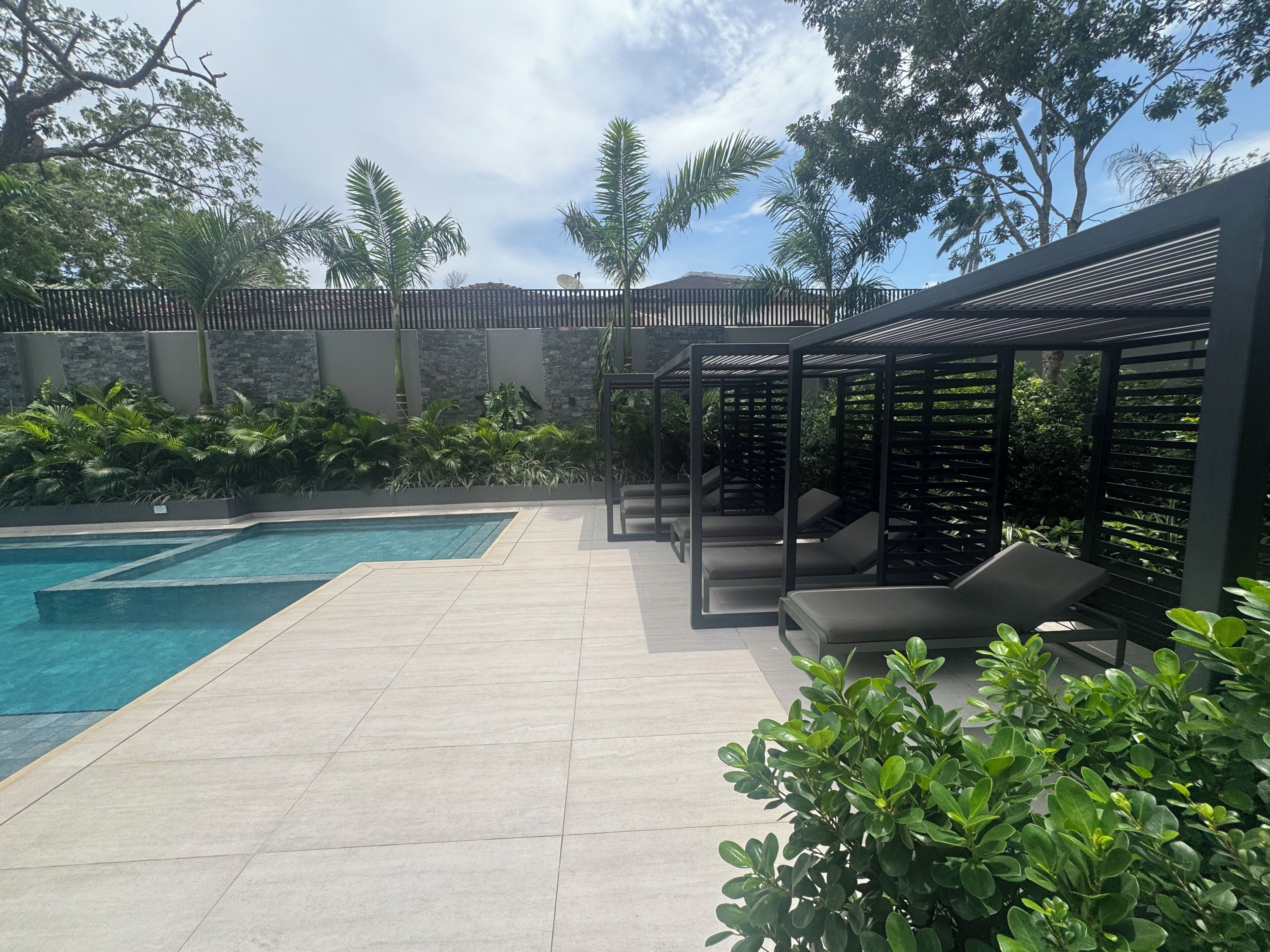 A swimming pool with a tiled deck, surrounded by greenery and a pergola with seating.