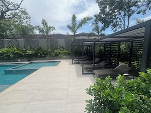 A swimming pool with a tiled deck, surrounded by greenery and a pergola with seating.