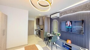 A modern living room with a flat-screen TV, a dining area with two chairs, a kitchen in the background, and a decorative wall panel. - Bantree Apartment by Cozy (Accra)