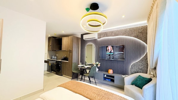 A modern hotel room with a bed, a dining area, a kitchen, and a TV mounted on the wall.