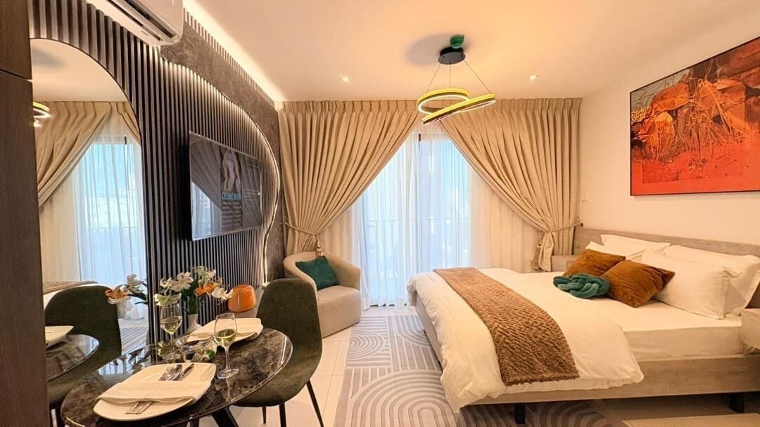 A modern hotel room with a bed, a dining area with a table and chairs, a sofa, and a large window with curtains.
