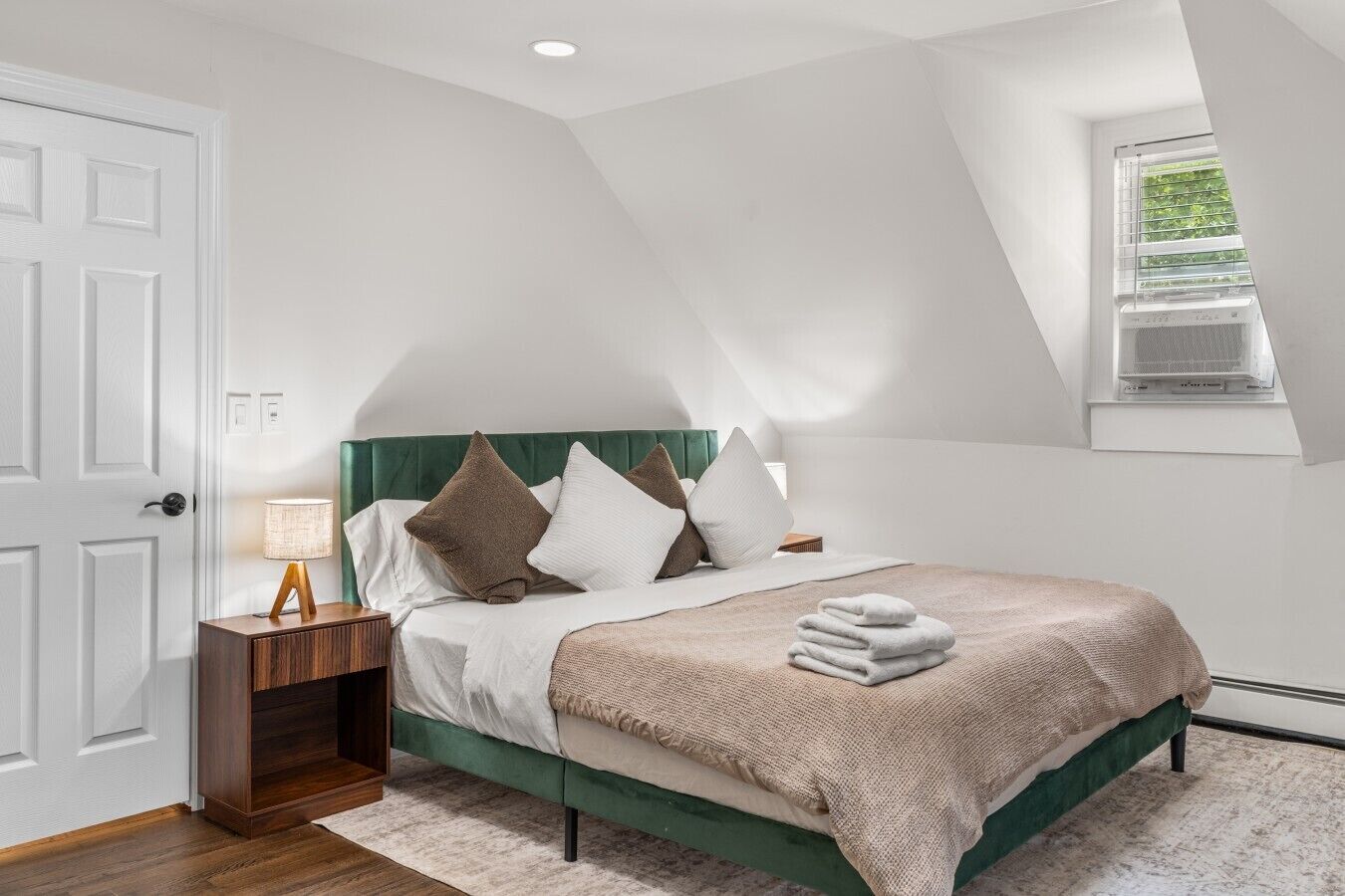 Un dormitorio con cama, almohadas, una mesita de noche, una lámpara, una ventana y una puerta.