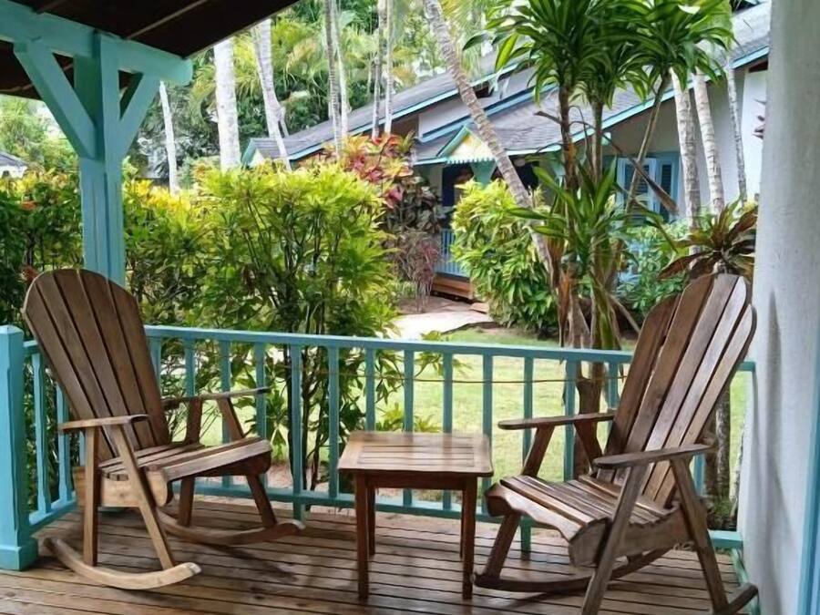 Una terraza de madera con mecedoras y una mesa, rodeada de exuberante follaje tropical y un edificio al fondo.