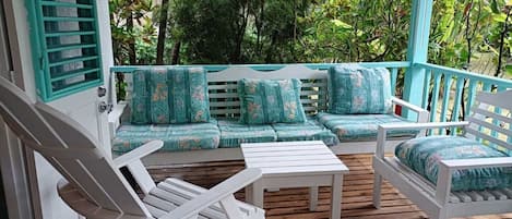 A wooden deck with a white sofa and chairs, surrounded by lush greenery.