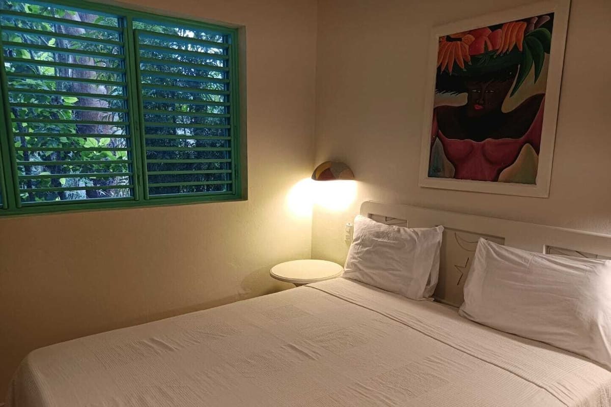Una cama con sábanas blancas, una mesita de noche con lámpara, una ventana con contraventanas verdes y un cuadro en la pared.